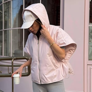 Puede incluir: Un chaleco con capucha rosa claro con cierre de cremallera y una gorra de béisbol blanca. El chaleco tiene un diseño único tipo capa y se combina con pantalones de chándal grises. Una persona sostiene una taza de café blanca.