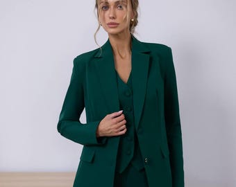 May include: A three-piece emerald green suit. The suit includes a blazer, a vest, and matching trousers. The blazer has a classic notched lapel and two-button closure. The vest is buttoned up. The suit is made of a smooth, solid fabric.