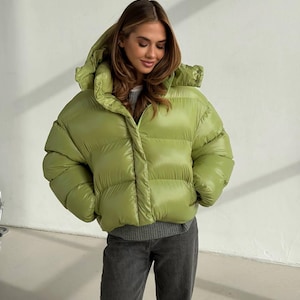 May include: A lime green puffer jacket with a glossy finish, featuring a high collar, a hood, and horizontal quilted channels. The jacket is worn over a grey jumper and dark grey jeans, showcasing a voluminous silhouette and snap closures.