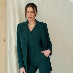 May include: A woman wearing a dark green suit with a vest and a gold necklace. The suit has a blazer with a notched lapel and a button closure.