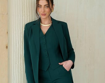 May include: A woman wearing a dark green suit with a vest and a gold necklace. The suit has a blazer with a notched lapel and a button closure.