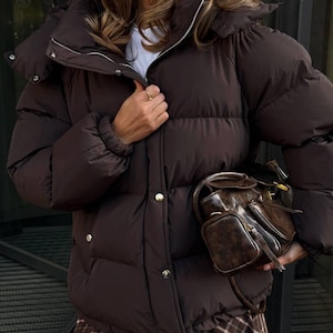 Puede incluir: Una chaqueta acolchada marrón oscuro con cuello alto y botones dorados. La chaqueta tiene un diseño acolchado y cierre de cremallera. Un bolso de mano de cuero marrón con múltiples compartimentos se sostiene en la mano.