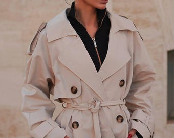 May include: Beige trench coat with a double-breasted design, featuring large brown buttons and a matching belt. The coat has a classic collar, shoulder epaulettes, and a black zipper visible underneath. A stylish outerwear piece.