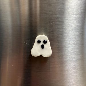 以下が含まれることがあります： 滑らかで光沢のある仕上げの、小さな白い幽霊の形をしたマグネット。この幽霊には2つの丸い黒い目と小さな楕円形の黒い口があり、丸みを帯びた不規則な形の体に、シンプルで驚いたような表情を作り出しています。