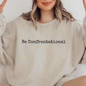 May include: A light beige sweatshirt with the text "Be Confrontational" printed in black on the front.