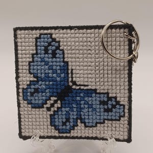 May include: A blue butterfly embroidered on a white canvas with a black border. The canvas is attached to a silver key ring.