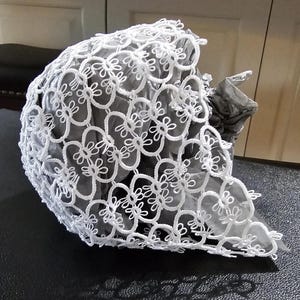 Puede incluir: Sombrero de encaje blanco tejido a crochet con un delicado patrón floral. El sombrero está hecho de un material fino y ligero y tiene un drapeado suave y fluido.