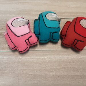 May include: Three plush Among Us crewmate toys in pink, teal, and red. Each character has a white visor and black outline. The toys are made of soft fabric and are designed to resemble the popular video game characters.