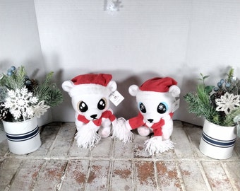 May include: Two white plush polar bears wearing red Santa hats and scarves. The bears have large eyes and are sitting on a brick surface. Decorative holiday greenery in white metal containers with blue stripes are on either side of the bears.