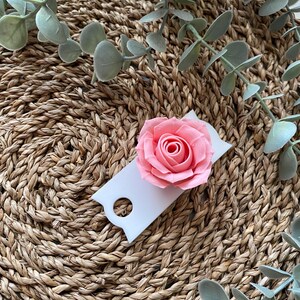 May include: A white tag with a hole in the top, featuring a pink paper rose. The tag is sitting on a woven straw mat with green eucalyptus leaves.