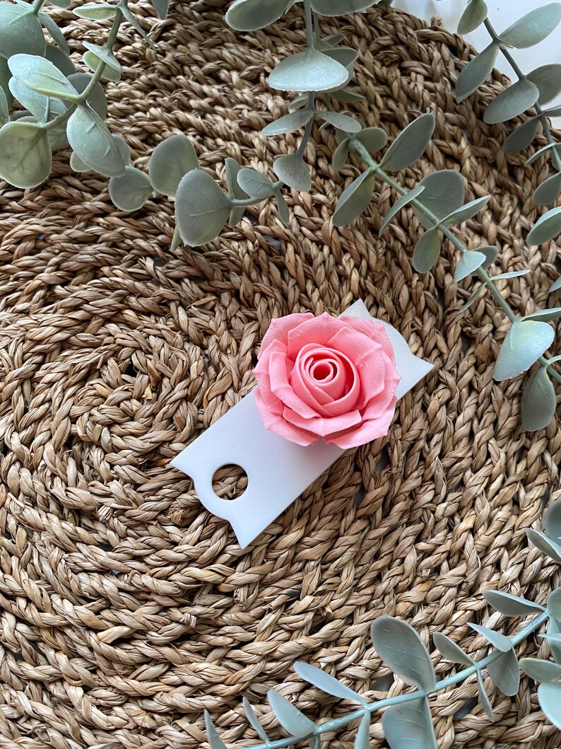 May include: A white tag with a hole in the top, featuring a pink paper rose. The tag is sitting on a woven straw mat with green eucalyptus leaves.