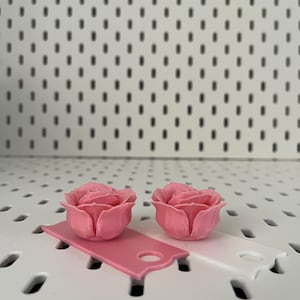 May include: Two pink 3D printed rose flower embellishments. The roses are on a white surface with small holes.