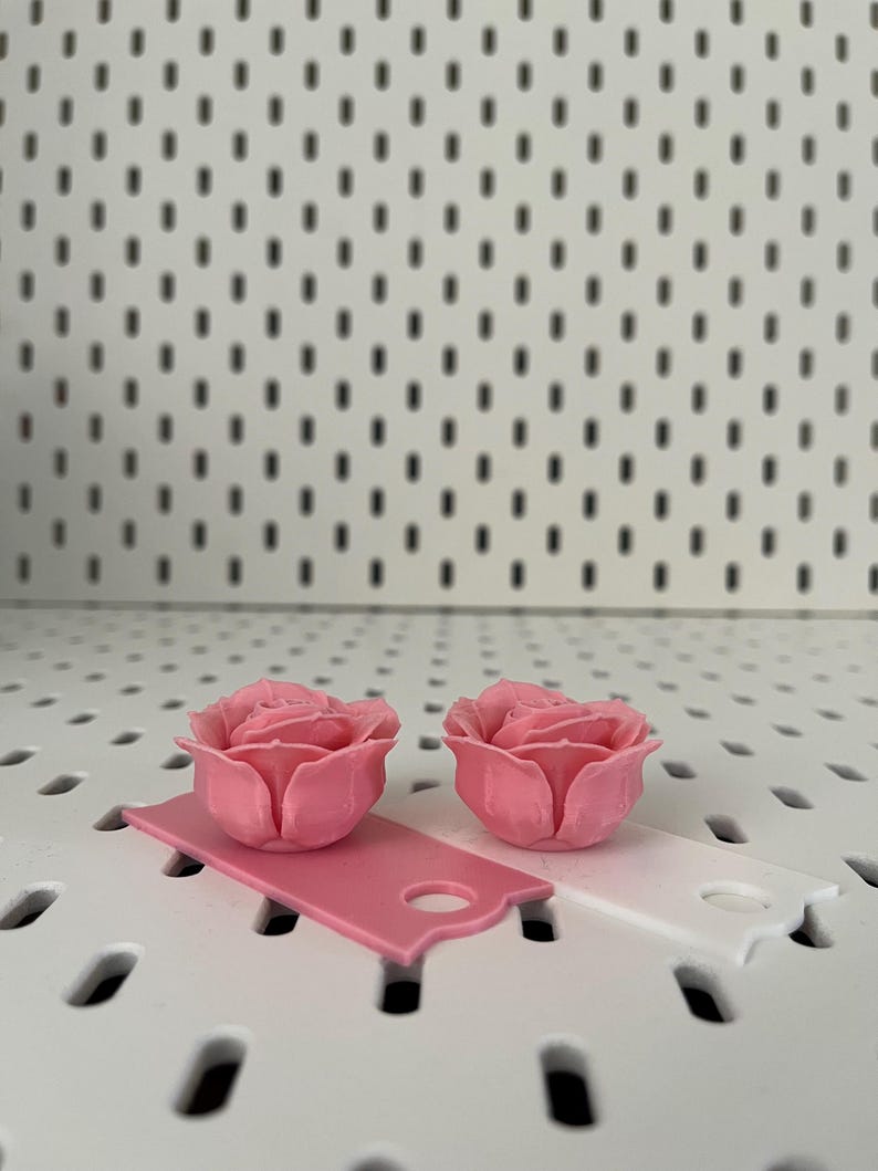 May include: Two pink 3D printed rose flower embellishments. The roses are on a white surface with small holes.