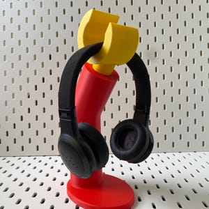 以下が含まれることがあります： 赤と黄色の3Dプリントされたヘッドホンスタンドに、黒いヘッドホンが置かれています。スタンドは曲線的なデザインで、円形のベースが付いています。ヘッドホンは黒いヘッドバンドとイヤーカップが付いています。