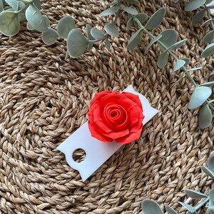 May include: A single red rose on a white tag with a hole in the center. The tag is on a woven brown surface with green leaves.