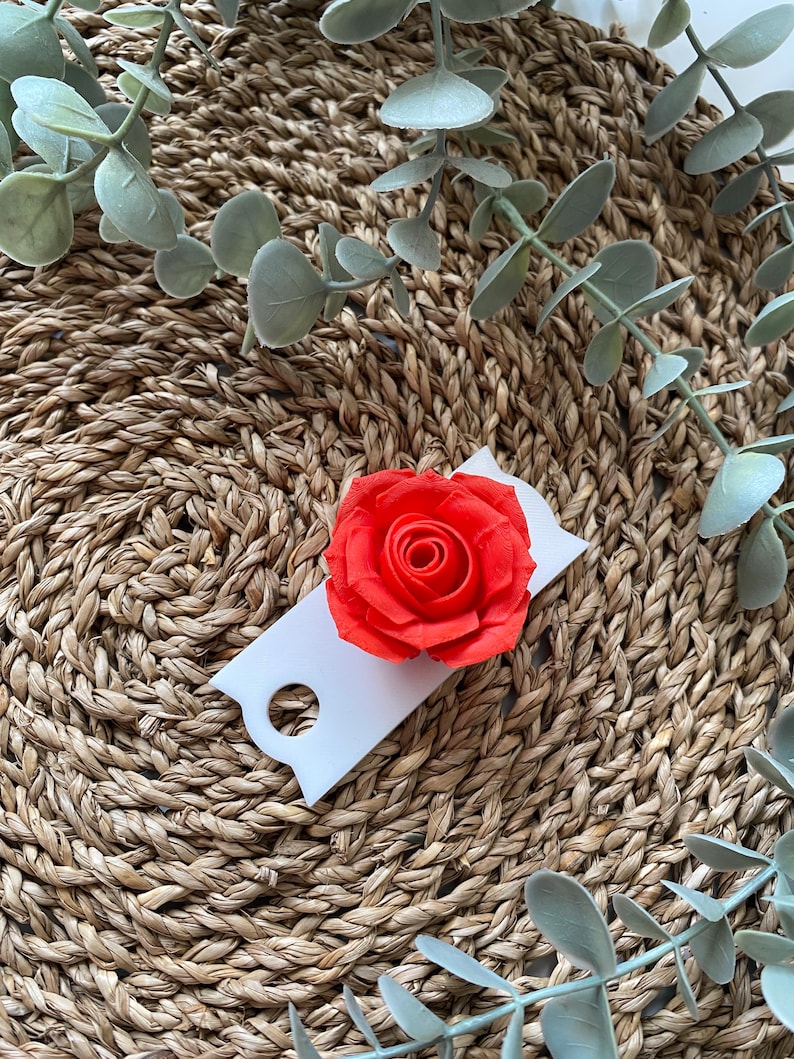 May include: A single red rose on a white tag with a hole in the center. The tag is on a woven brown surface with green leaves.