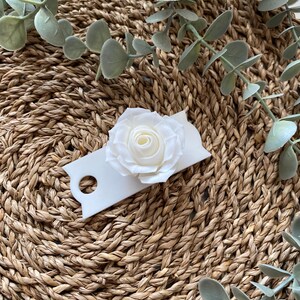 May include: A white paper tag with a white paper rose on top. The tag has a hole punched in the top left corner. The tag is sitting on a woven brown surface.