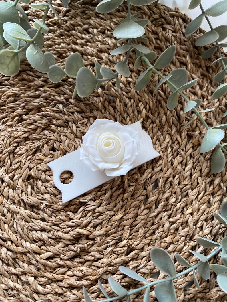May include: A white paper tag with a white paper rose on top. The tag has a hole punched in the top left corner. The tag is sitting on a woven brown surface.