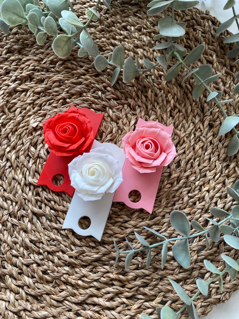 May include: Three paper rose flower embellishments on a woven background. The roses are red, pink, and white. Each rose is attached to a small rectangular tag.