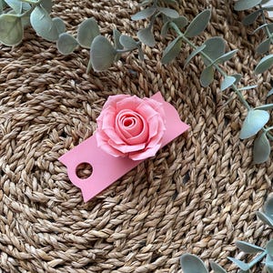 May include: A pink paper tag with a hole in the top and a pink paper rose attached to the front. The tag is sitting on a woven straw surface.