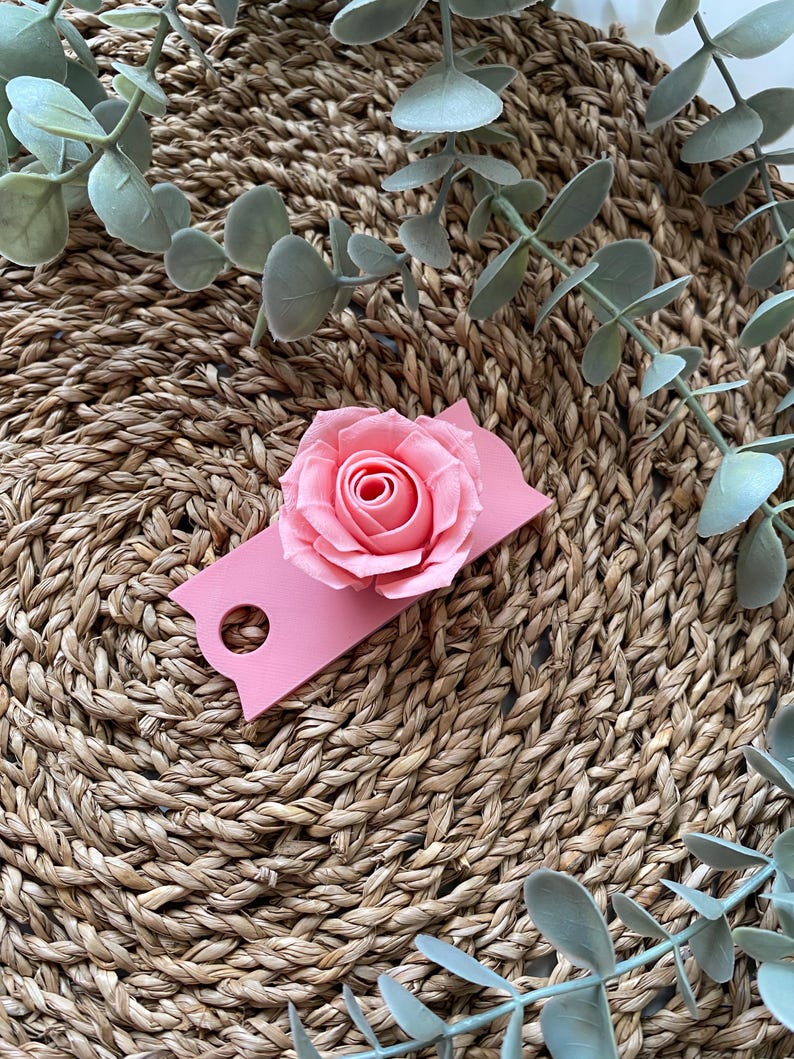 May include: A pink paper tag with a hole in the top and a pink paper rose attached to the front. The tag is sitting on a woven straw surface.