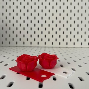 May include: Two red 3D printed rose flower embellishments on a white pegboard background. The roses are on top of two small red and white rectangular pieces of paper.