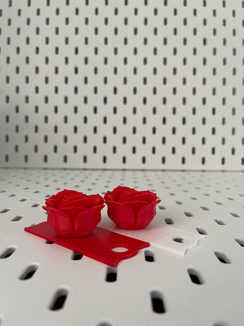 May include: Two red 3D printed rose flower embellishments on a white pegboard background. The roses are on top of two small red and white rectangular pieces of paper.