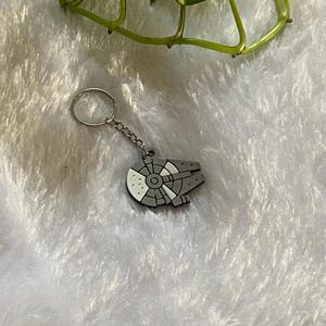 May include: A gray Millennium Falcon keychain with white and gray details. The keychain has a silver chain and ring. The background is a white, fluffy surface with a green plant in the upper right corner.