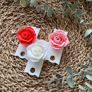 May include: Three white ceramic flower holders with a scalloped edge and a hole in the center. Each holder features a different colored paper rose: red, pink, and white.
