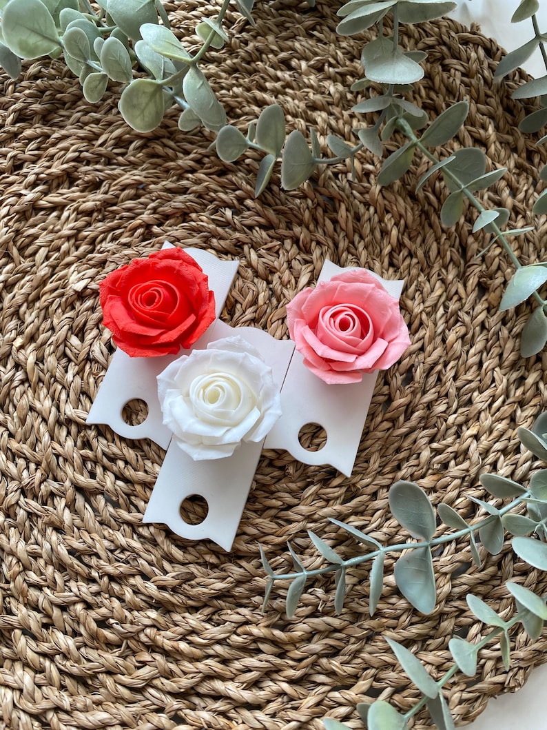 May include: Three white ceramic flower holders with a scalloped edge and a hole in the center. Each holder features a different colored paper rose: red, pink, and white.