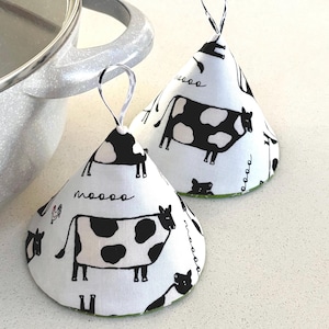 May include: Two white and black cow-print pot holders with a loop for hanging. The pot holders are cone-shaped and feature a repeating pattern of cows and the word "moooo". The edges are trimmed with green fabric.