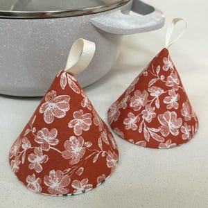 May include: Two orange pot holders with a white floral pattern and white ribbon ties. The pot holders are shaped like cones.