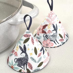 May include: Two cone-shaped pot holders with a floral and bunny print. The pot holders have a white background with pink, green, and blue floral and leaf patterns. Each pot holder has a navy blue loop for hanging.