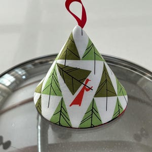 May include: A cone-shaped fabric pot lid handle cover featuring a white background with a repeating pattern of green pine trees and a stylized red figure carrying a tree. The cover is topped with a small red fabric loop for hanging.