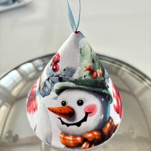 May include: A cone-shaped fabric ornament featuring a printed snowman with a carrot nose, a green hat, and an orange plaid scarf. The ornament has a light blue ribbon loop at the top for hanging and displays red berries and green foliage accents.
