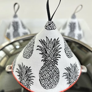 May include: A white pot holder with a black pineapple pattern and a black loop handle. The pot holder is on a black surface.