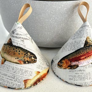 May include: Two white fabric pot holders with a brown and orange fish design. The pot holders are shaped like cones and have a loop handle.