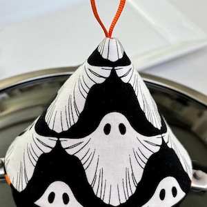 May include: A cone-shaped fabric pot handle cover featuring a repeating black and white ghost pattern. The textile accessory includes a bright orange ribbon loop at the top for hanging and small orange accents along the bottom edge of the cover.