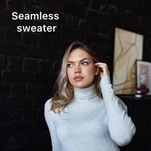 Peut inclure: Une femme portant un pull col roulé gris clair sans couture et un jean bleu. Elle se tient devant un mur de briques noires.