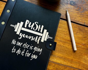 May include: Black leather journal with a gold metal clasp and a white inscription that reads "PUSH yourself No one else is going to do it for you". The journal is on a wooden table with a cup of coffee and a wooden pen.