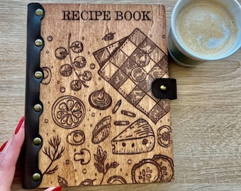Puede incluir: Un libro de recetas de madera con una cubierta de cuero negro y un diseño de varios alimentos, incluyendo queso, verduras y hierbas. El libro tiene las palabras "RECIPE BOOK" grabadas en la portada.