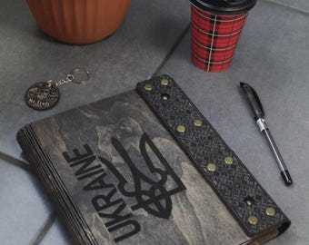 May include: A black wooden notebook with a leather strap and gold accents. The notebook has the word "UKRAINE" and a trident symbol engraved on the front.