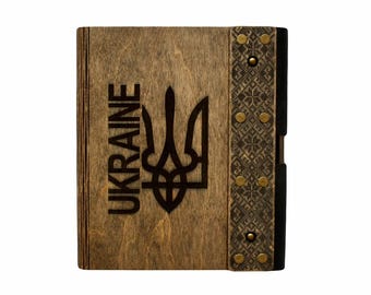 May include: A wooden journal with a dark brown finish. The cover features a black Ukrainian trident and the word "UKRAINE" in black lettering. The spine is decorated with a black and white geometric pattern and has brass accents.