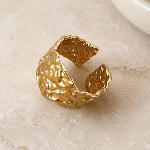 May include: A gold-toned open ring featuring a wide, irregular band with a highly textured, hammered surface. The metal has a reflective, polished finish with deep indentations across the entire exterior, creating a wavy, organic edge profile.
