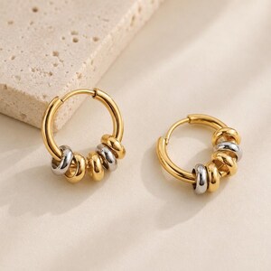 May include: A pair of hoop earrings featuring a polished gold-tone circular frame. Each hoop is adorned with a series of small, alternating gold and silver-tone metallic rings that slide freely along the main hoop structure.