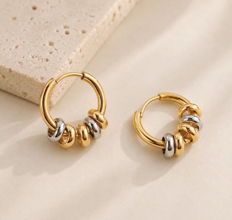 May include: A pair of hoop earrings featuring a polished gold-tone circular frame. Each hoop is adorned with a series of small, alternating gold and silver-tone metallic rings that slide freely along the main hoop structure.