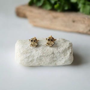 May include: A pair of gold-colored stud earrings shaped like a character with large ears and dark eyes. The earrings are displayed on a white, textured stone, with a blurred background of greenery and wood.