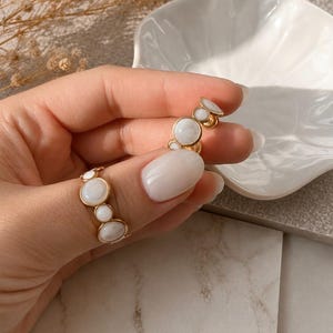 May include: A gold-toned ring featuring a series of circular, milky white cabochon stones set in a continuous band. The stones vary slightly in size, alternating between larger and smaller round white gems held within a polished gold metal frame.