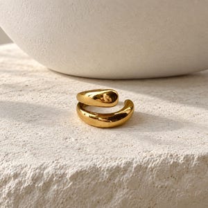 May include: A polished gold-tone ring featuring an open, bypass-style design with two smooth, rounded ends that overlap slightly. The metal surface is highly reflective, catching the light to highlight its curved, fluid, and continuous shape.