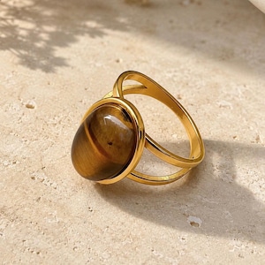 May include: A gold-toned ring featuring a smooth, oval-shaped tiger's eye gemstone set in a polished metal bezel. The band consists of two thin, parallel metal wires that meet at the base of the setting, creating an open, layered design.